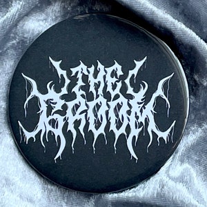 May include: A black and white button with the text "THE GHOUL" in a stylized, gothic font.