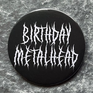 May include: A black and white button with the text "Birthday Metalhead" in a stylized metal font.