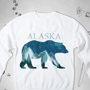 Puede incluir: Sudadera blanca con un gr&aacute;fico de silueta de oso y cordillera de monta&ntilde;as azules y verdes. El texto "ALASKA" est&aacute; impreso sobre el gr&aacute;fico.