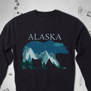 Puede incluir: Una sudadera azul oscuro con un gr&aacute;fico de una silueta de oso con monta&ntilde;as y estrellas en su interior. El texto "ALASKA" est&aacute; impreso sobre el oso.