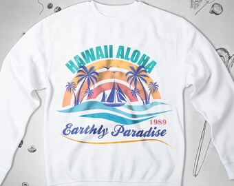 Op de afbeelding: Witte trui met een kleurrijk grafisch ontwerp met palmbomen, een zeilboot en de tekst "Hawaii Aloha Earthly Paradise 1989".