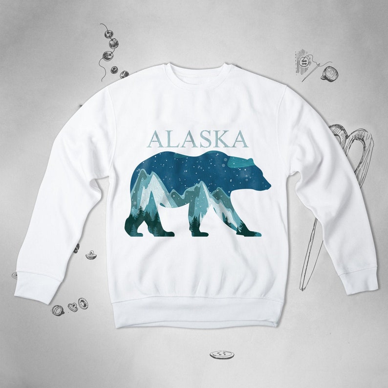 Puede incluir: Sudadera blanca con un gr&aacute;fico de silueta de oso y cordillera de monta&ntilde;as azules y verdes. El texto "ALASKA" est&aacute; impreso sobre el gr&aacute;fico.