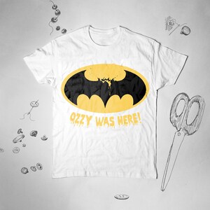 May include: White t-shirt with a yellow Batman logo and the text "Ozzy Was Here!" in dripping yellow letters.