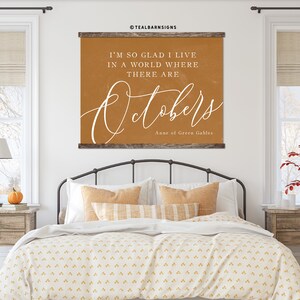 May include: A framed wall art piece with the text "I'M SO GLAD I LIVE IN A WORLD WHERE THERE ARE Octobers" in white script on an orange background. The artwork is from Anne of Green Gables and is displayed above a bed.