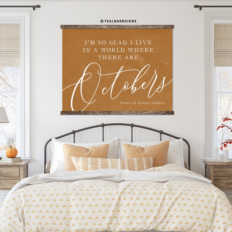 May include: A framed wall art piece with the text "I'M SO GLAD I LIVE IN A WORLD WHERE THERE ARE Octobers" in white script on an orange background. The artwork is from Anne of Green Gables and is displayed above a bed.