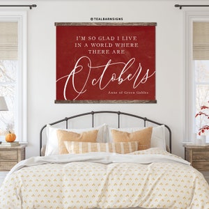 May include: A red wall hanging with the text "I'M SO GLAD I LIVE IN A WORLD WHERE THERE ARE Octobers" in white script. The sign is framed with a dark wood border and is displayed above a bed.
