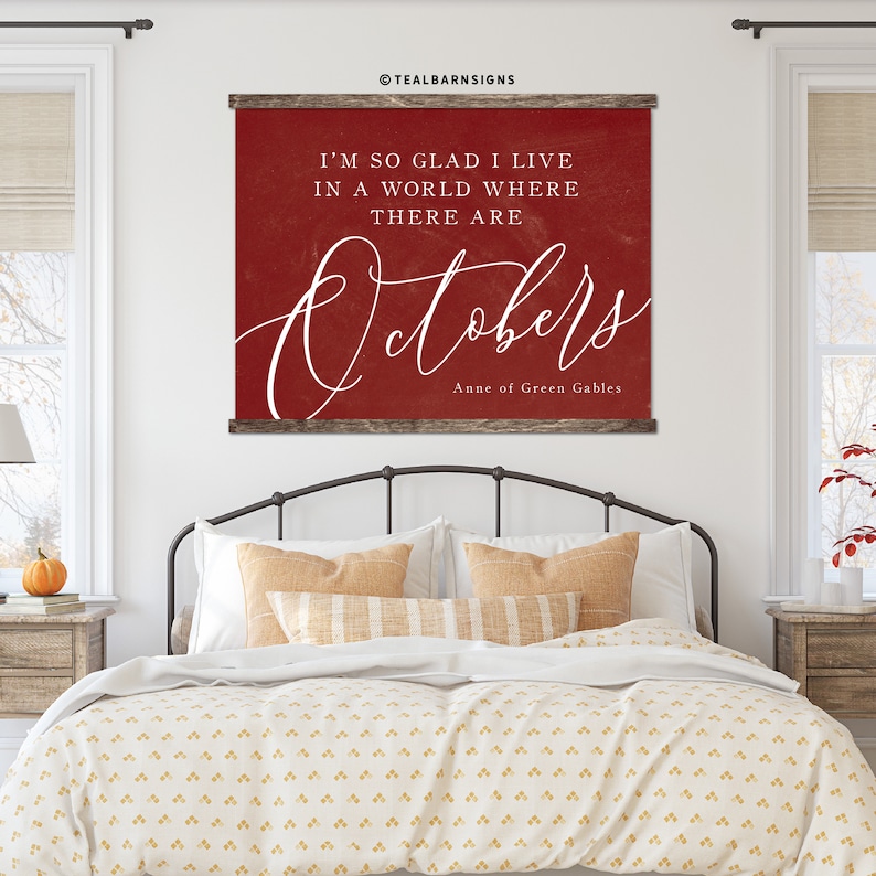 May include: A red wall hanging with the text "I'M SO GLAD I LIVE IN A WORLD WHERE THERE ARE Octobers" in white script. The sign is framed with a dark wood border and is displayed above a bed.