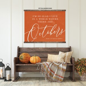 May include: An orange wall hanging with the text "I'M SO GLAD I LIVE IN A WORLD WHERE THERE ARE Octobers" and "Anne of Green Gables". The image also features a wooden bench, pumpkins, pillows, a plaid blanket, and lanterns.