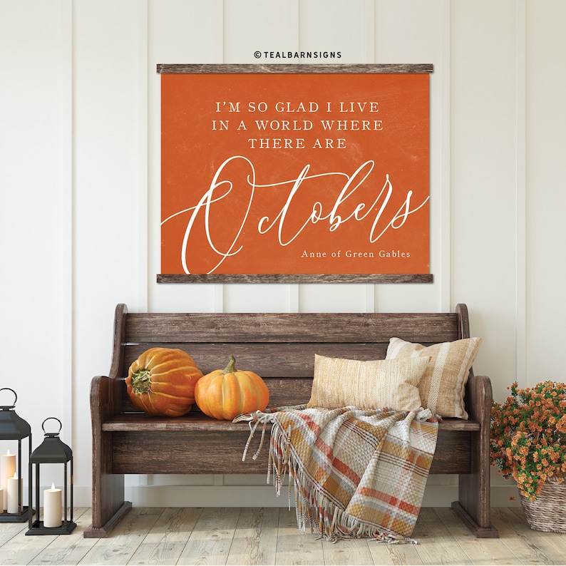 May include: An orange wall hanging with the text "I'M SO GLAD I LIVE IN A WORLD WHERE THERE ARE Octobers" and "Anne of Green Gables". The image also features a wooden bench, pumpkins, pillows, a plaid blanket, and lanterns.