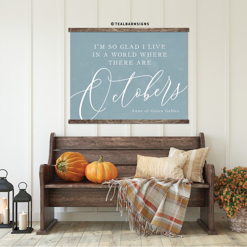 May include: A wooden bench with two pumpkins, pillows, and a plaid blanket. Above the bench is a blue sign with the text "I'M SO GLAD I LIVE IN A WORLD WHERE THERE ARE Octobers" and "Anne of Green Gables".
