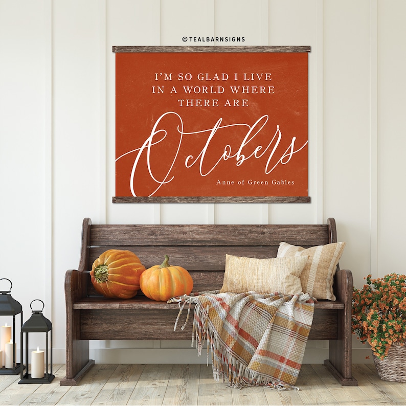 May include: A rustic wooden bench is adorned with pumpkins, pillows, and a plaid throw. Above, a sign with the text "I'M SO GLAD I LIVE IN A WORLD WHERE THERE ARE Octobers" adds a touch of autumn charm. The sign is from Anne of Green Gables.