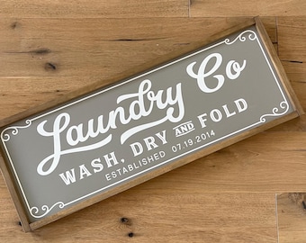 May include: A wooden sign with a gray background and white text that reads "Laundry Co. Wash, Dry and Fold Established 07.19.2014". The sign has a decorative border with a scroll design.
