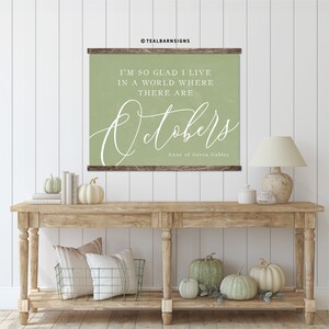 May include: A framed wall art piece with the text "I'm so glad I live in a world where there are Octobers" in white script on a sage green background. The artwork is displayed above a wooden console table decorated with pumpkins and fall decor.