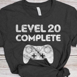 May include: A dark grey t-shirt with the text "Level 20 Complete" in white. A white video game controller graphic is below the text.
