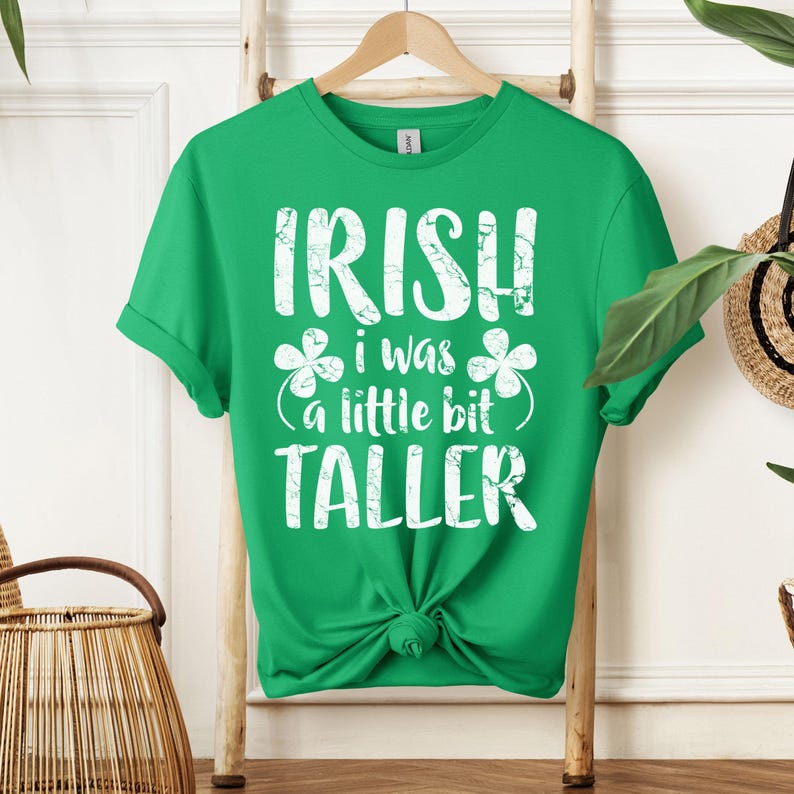 K&ouml;nnte beinhalten: Gr&uuml;nes T-Shirt mit wei&szlig;em Text, der "IRISH I WAS A LITTLE BIT TALLER" lautet, mit Kleeblatt-Designs auf beiden Seiten des Textes.