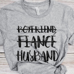 May include: Gray t-shirt with black text that reads "Boyfriend, Fiance, Husband" with lines through the first two words.