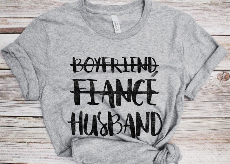 May include: Gray t-shirt with black text that reads "Boyfriend, Fiance, Husband" with lines through the first two words.