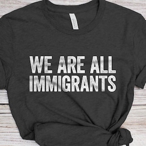 May include: Dark gray t-shirt with the white text "WE ARE ALL IMMIGRANTS". The shirt is made of a soft material and has a crew neck. The text is in a bold, sans-serif font.