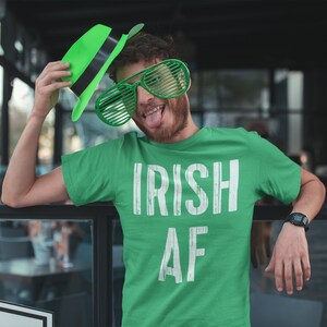 May include: A green t-shirt with the text "IRISH AF" in white. The person wearing the shirt is holding a green hat and wearing green oversized sunglasses.
