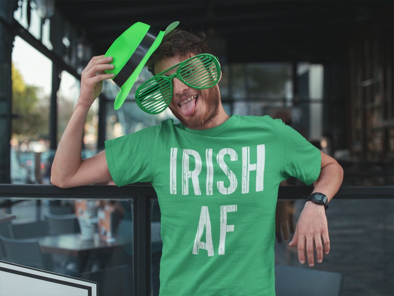 May include: A green t-shirt with the text "IRISH AF" in white. The person wearing the shirt is holding a green hat and wearing green oversized sunglasses.