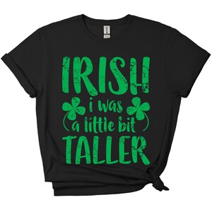 K&ouml;nnte beinhalten: Schwarzes T-Shirt mit einem gr&uuml;nen, abgenutzten Aufdruck, der "Irish I was a little bit taller" lautet, mit Kleeblatt-Designs auf beiden Seiten des Textes.