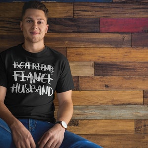 May include: A black t-shirt with white text that reads "Boyfriend Fiance Husband" on a man wearing blue jeans against a wood background.
