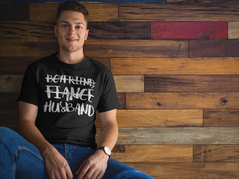 May include: A black t-shirt with white text that reads "Boyfriend Fiance Husband" on a man wearing blue jeans against a wood background.