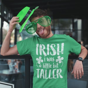 K&ouml;nnte beinhalten: Ein gr&uuml;nes T-Shirt mit dem Text "IRISH I WAS A LITTLE BIT TALLER" in Wei&szlig; mit einem Kleeblatt auf jeder Seite des Textes. Die Person, die das Shirt tr&auml;gt, h&auml;lt einen gr&uuml;nen Hut und tr&auml;gt eine gr&uuml;ne Sonnenbrille.