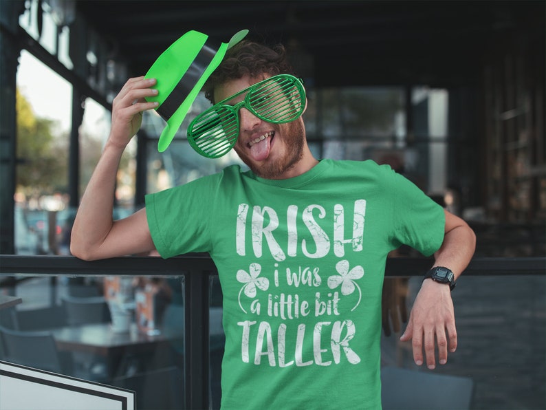 K&ouml;nnte beinhalten: Ein gr&uuml;nes T-Shirt mit dem Text "IRISH I WAS A LITTLE BIT TALLER" in Wei&szlig; mit einem Kleeblatt auf jeder Seite des Textes. Die Person, die das Shirt tr&auml;gt, h&auml;lt einen gr&uuml;nen Hut und tr&auml;gt eine gr&uuml;ne Sonnenbrille.