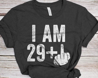 May include: A dark gray t-shirt with white text that reads "I AM 29+" and a white hand giving the middle finger.