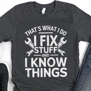 Peut inclure: Un t-shirt gris foncé avec un motif blanc qui dit "That's what I do I fix stuff and I know things". Le motif comprend également deux clés à molette croisées.