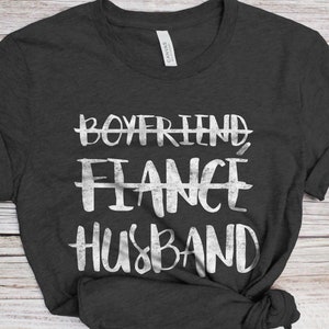 May include: A dark gray t-shirt with a white graphic that says "Boyfriend, Fiance, Husband" with lines through the first two words.