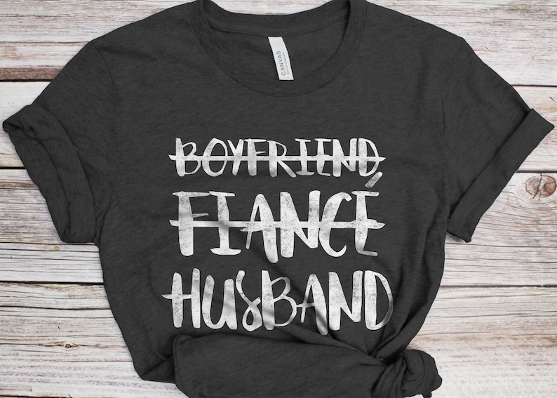 May include: A dark gray t-shirt with a white graphic that says "Boyfriend, Fiance, Husband" with lines through the first two words.