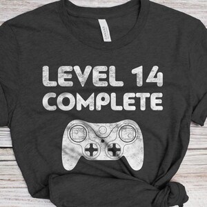 May include: A dark gray t-shirt with the text "LEVEL 14 COMPLETE" in white, and a white graphic of a video game controller.
