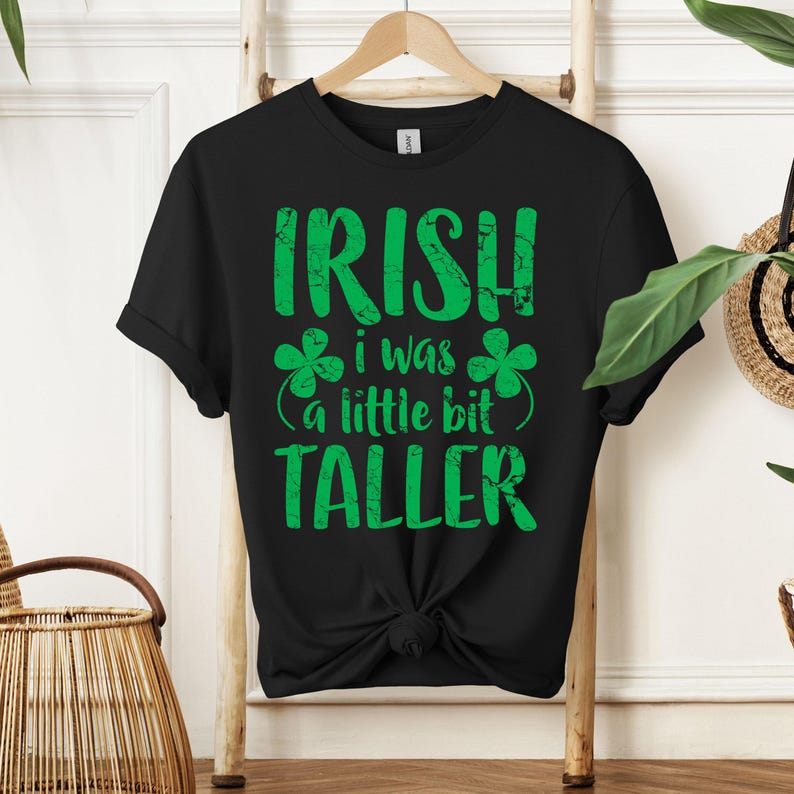 K&ouml;nnte beinhalten: Schwarzes T-Shirt mit einer gr&uuml;nen, abgenutzten Grafik, auf der "IRISH I WAS A LITTLE BIT TALLER" mit Kleeblatt-Designs steht.