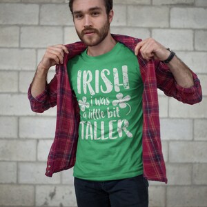 K&ouml;nnte beinhalten: Ein Mann tr&auml;gt ein gr&uuml;nes T-Shirt mit einem wei&szlig;en, abgenutzten Aufdruck, auf dem "IRISH I WAS A LITTLE BIT TALLER" steht, und ein rot-schwarzes Karohemd. Er steht vor einer wei&szlig;en Backsteinwand.