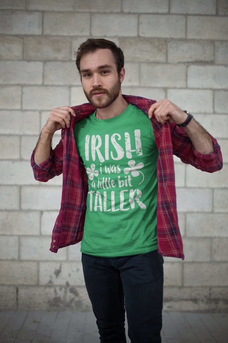 K&ouml;nnte beinhalten: Ein Mann tr&auml;gt ein gr&uuml;nes T-Shirt mit einem wei&szlig;en, abgenutzten Aufdruck, auf dem "IRISH I WAS A LITTLE BIT TALLER" steht, und ein rot-schwarzes Karohemd. Er steht vor einer wei&szlig;en Backsteinwand.