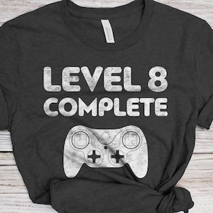 May include: A dark gray t-shirt with the text "Level 8 Complete" in white with a distressed look. A white video game controller graphic is below the text.
