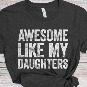 May include: A dark gray t-shirt with the text "Awesome Like My Daughters" printed in white.