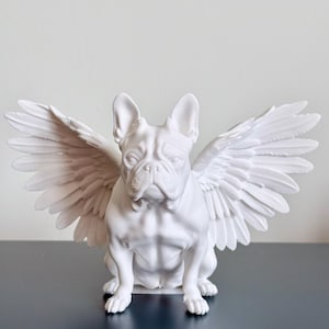 Pode incluir: Escultura em cerâmica branca de um buldogue francês com asas. O cão está sentado com as asas abertas. A escultura tem um acabamento liso e é um elemento decorativo.