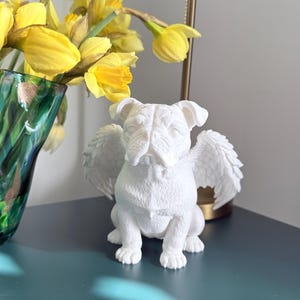 Könnte beinhalten: Eine weiße Bulldoggenfigur mit Engelsflügeln, die auf einer teal-farbenen Oberfläche sitzt. Die Skulptur befindet sich neben einer Vase mit gelben Narzissen. Im Hintergrund ist eine Lampe zu sehen.