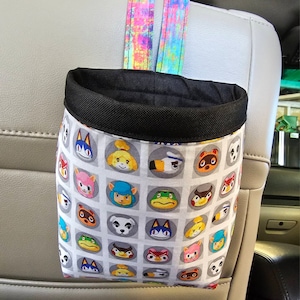 May include: A black fabric car trash can with a tie-top closure. The can is decorated with a repeating pattern of Animal Crossing characters in colorful circles on a gray background.