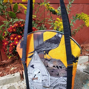 Op de afbeelding: Een zwarte tote bag met een ritssluiting en twee zwarte banden. De tas heeft een kleurrijke print van een spookhuis met een kraai die op het dak zit. Het huis heeft gebroken ramen en een lantaarn die aan de zijkant hangt.