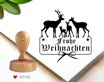 May include: A black and white rubber stamp with a design of a family of deer and the text "Frohe Weihnachten" in a decorative font. The stamp is mounted on a wooden base.