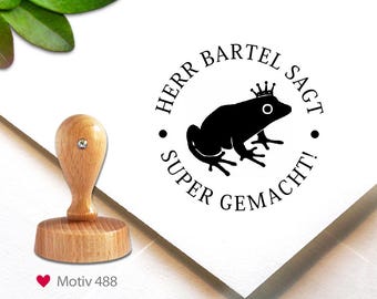 May include: A wooden stamp with a light brown handle and base, featuring a black frog wearing a crown inside a circular design. The text reads "HERR BARTEL SAGT SUPER GEMACHT!" and "Motiv 488".