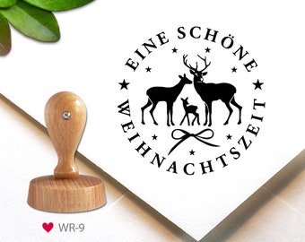 May include: A round wooden stamp with a black design of a deer family and the text "Eine schöne Weihnachtszeit" in a circle. The stamp is on a white background.