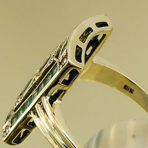 Pu&ograve; includere: Un anello d'oro con una faccia rettangolare allungata. La faccia presenta un motivo di pietre verde smeraldo e piccole pietre trasparenti. Il gambo dell'anello &egrave; in oro lucido semplice. L'anello &egrave; posto su uno sfondo neutro.