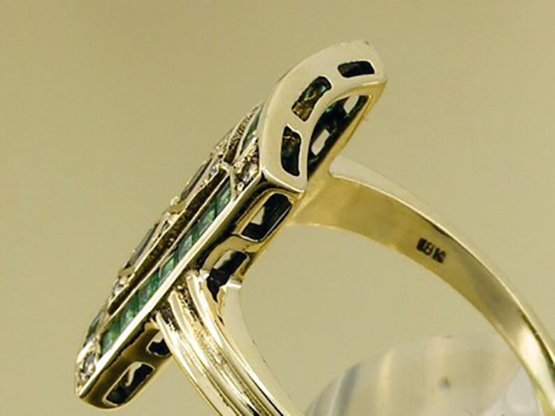 Pu&ograve; includere: Un anello d'oro con una faccia rettangolare allungata. La faccia presenta un motivo di pietre verde smeraldo e piccole pietre trasparenti. Il gambo dell'anello &egrave; in oro lucido semplice. L'anello &egrave; posto su uno sfondo neutro.