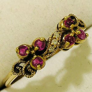 Op de afbeelding: Een goudkleurige ring met een bloemmotief, gezet met verschillende ronde, dieproze edelstenen en kleine, heldere accentstenen. De ring heeft een sierlijke, filigraan-achtige zetting en een delicate band.