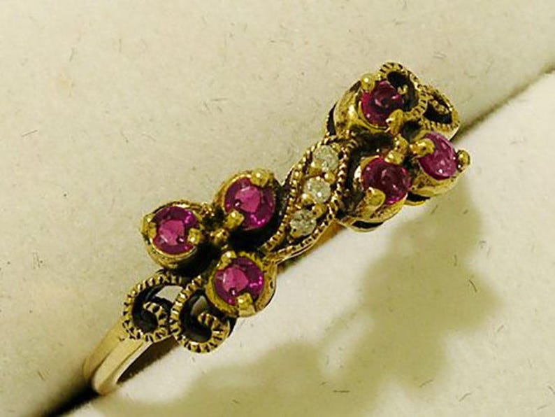Op de afbeelding: Een goudkleurige ring met een bloemmotief, gezet met verschillende ronde, dieproze edelstenen en kleine, heldere accentstenen. De ring heeft een sierlijke, filigraan-achtige zetting en een delicate band.